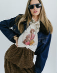 Woman wearing a denim jacket, graphic t-shirt, and brown pants with sunglasses on a white background