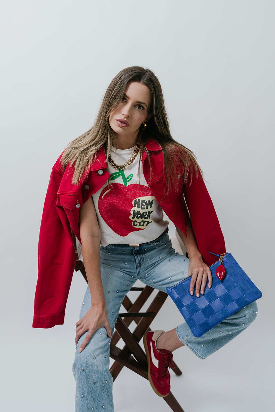 Woman wearing a red jacket and blue jeans, holding a blue handbag with a red heart, on a white background.