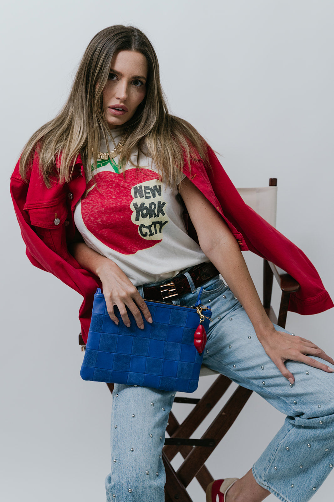 Woman wearing a red jacket, white t-shirt with graphic design, and blue jeans, holding a blue bag.