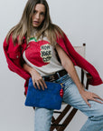 Woman wearing a red jacket, white t-shirt with graphic design, and blue jeans, holding a blue bag.