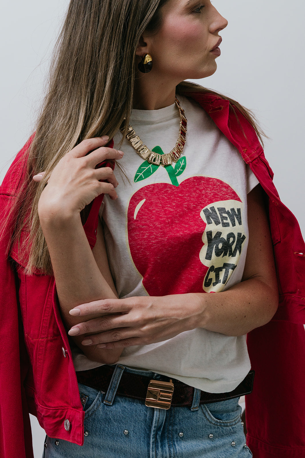 Woman wearing a white t-shirt with a red apple graphic and 'New York City' text, paired with a red jacket, close up front view