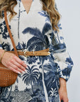Woman wearing a blue and white patterned dress with a belt, holding a brown bag against a white background.