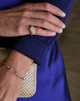 A close-up lifestyle shot of a person's hand wearing a gold ring with a central clear gemstone, paired with a pearl-embellished clutch and a gold bracelet against a royal blue dress.