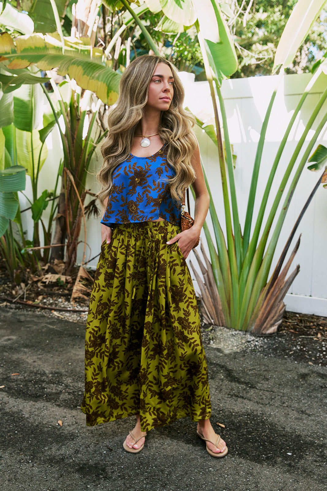 A woman stands outdoors in a tropical garden setting, wearing a sleeveless top with a blue and brown floral pattern and a long, wide-leg olive green skirt with a dark brown leaf print. She is wearing tan sandals and a necklace.