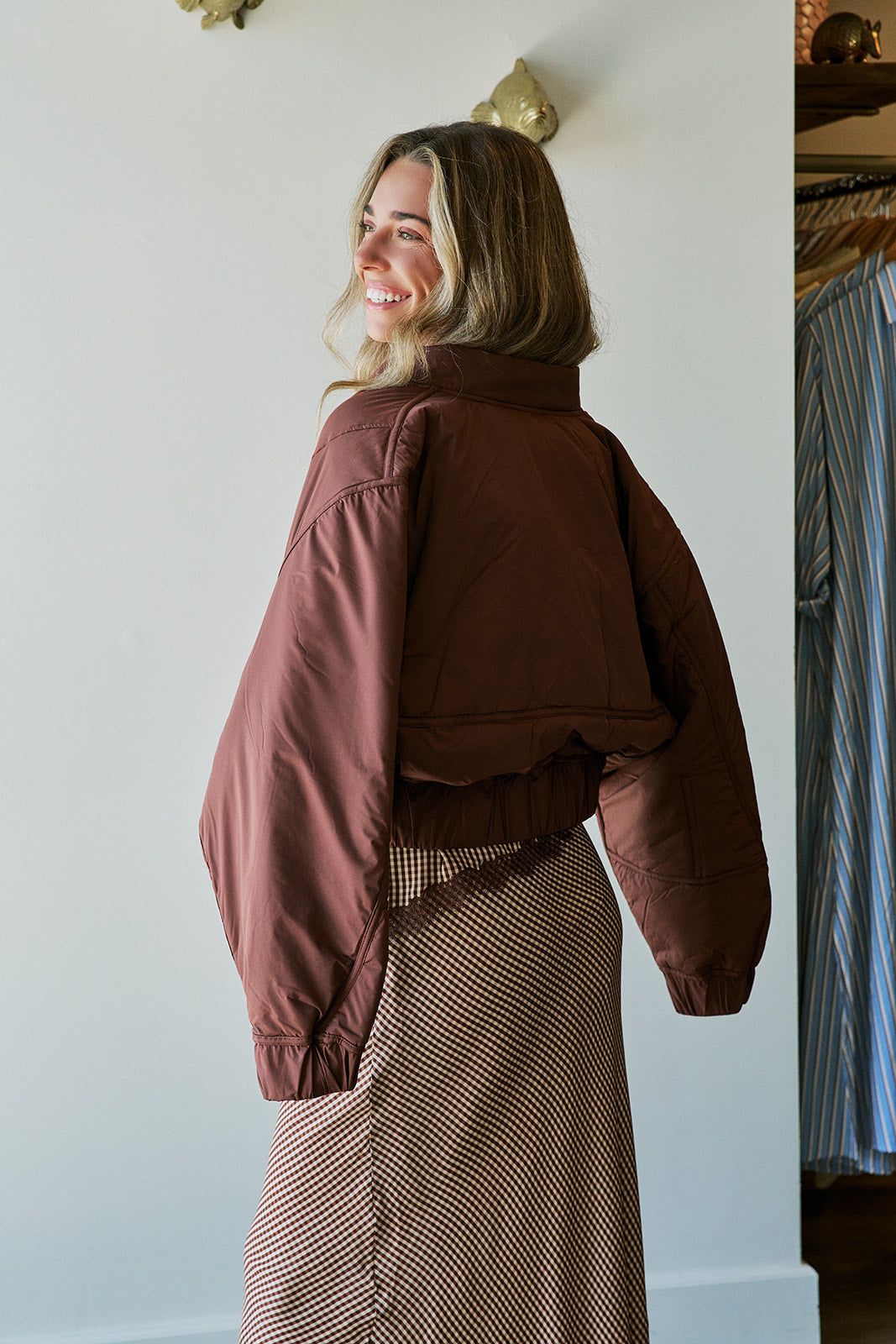A woman wearing a cropped, oversized brown quilted jacket and a brown-and-white patterned skirt, photographed indoors against a white wall. Back view