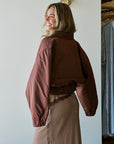 A woman wearing a cropped, oversized brown quilted jacket and a brown-and-white patterned skirt, photographed indoors against a white wall. Back view
