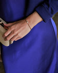 A close-up lifestyle shot of a person wearing a royal blue dress and a textured gold clutch, showcasing a delicate gold chain bracelet with a small clear crystal pendant on their wrist.
