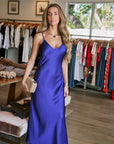 A woman modeling a royal blue satin slip dress inside a boutique clothing store, holding a gold clutch.
