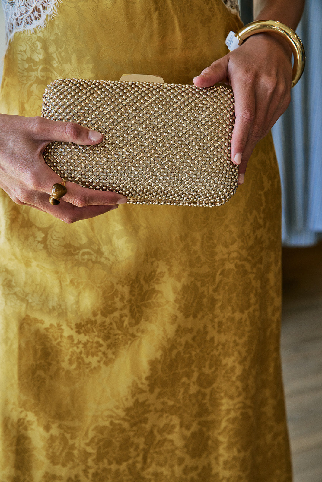 A close-up of a person holding a structured clutch bag covered in small metallic gold beads. The person is wearing a textured gold dress and gold jewelry, including a chunky gold bangle and a ring.