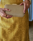 A close-up of a person holding a structured clutch bag covered in small metallic gold beads. The person is wearing a textured gold dress and gold jewelry, including a chunky gold bangle and a ring.