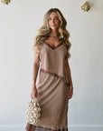 A woman models a brown and beige gingham slip set with dark brown lace trim, holding beaded bag against a white wall decorated with gold wall sconces.