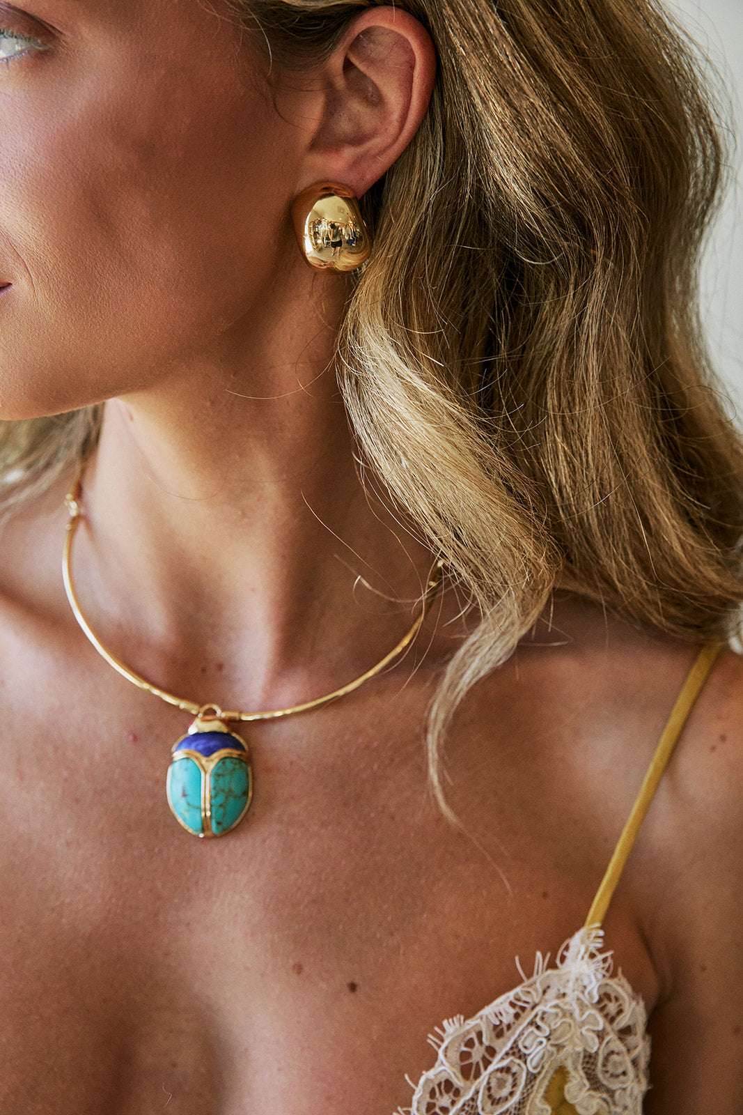 A close-up lifestyle shot of a woman wearing a gold necklace with a turquoise and gold scarab beetle pendant, along with a matching gold earring.