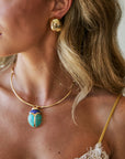 A close-up lifestyle shot of a woman wearing a gold necklace with a turquoise and gold scarab beetle pendant, along with a matching gold earring.