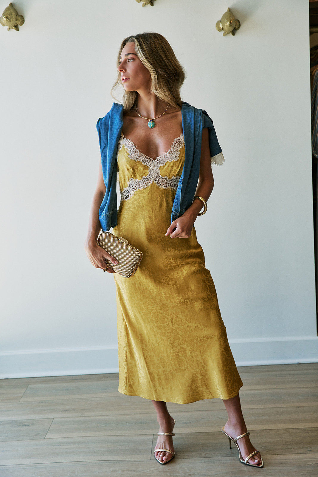 A woman models a yellow midi slip dress with a lace-trimmed neckline, styled with a blue shirt over her shoulders, a gold clutch, and gold high-heeled sandals against a white wall.