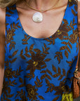 A close-up of a person wearing a bright blue sleeveless top with a brown floral and leaf pattern, a gold necklace with a large white shell pendant, and carrying a brown shoulder bag.