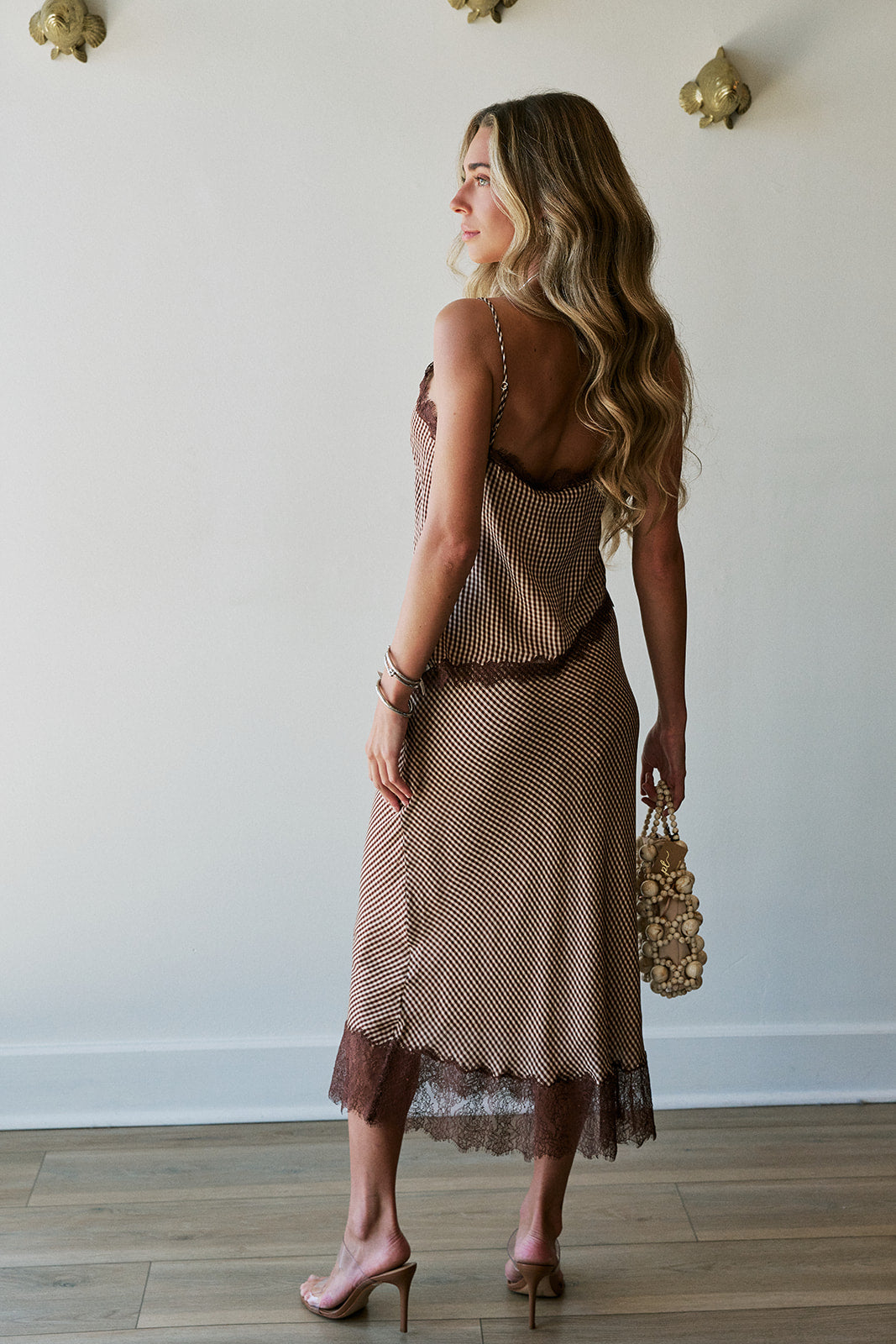 A woman stands in profile wearing a brown and beige set with lace trim, holding a woven clutch bag, set against a minimalist white wall with gold wall sconces. Back view