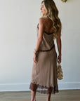 A woman stands in profile wearing a brown and beige set with lace trim, holding a woven clutch bag, set against a minimalist white wall with gold wall sconces. Back view