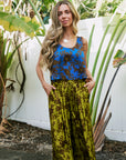 A woman stands outdoors in front of a white fence and tropical plants, wearing a sleeveless blue and brown floral top with olive green and brown patterned wide-leg pants with Ciao Bella Mother of Pearl Bar Choker Necklace