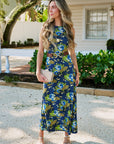 A woman stands outdoors in front of a house porch wearing a sleeveless navy maxi dress with a blue, green, and yellow floral print, a brown belt with gold hardware, and holding a beige clutch.