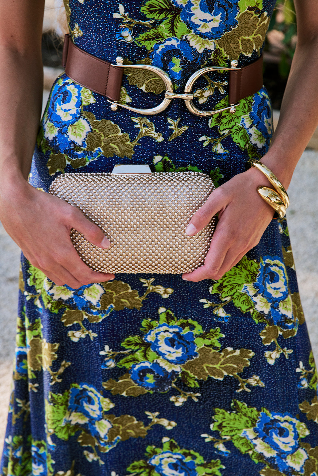 A close-up lifestyle shot of a person wearing a navy blue floral print dress with blue, green, and gold flowers, a brown belt with a large gold buckle, and holding Natasha Ball Mesh Clutch
