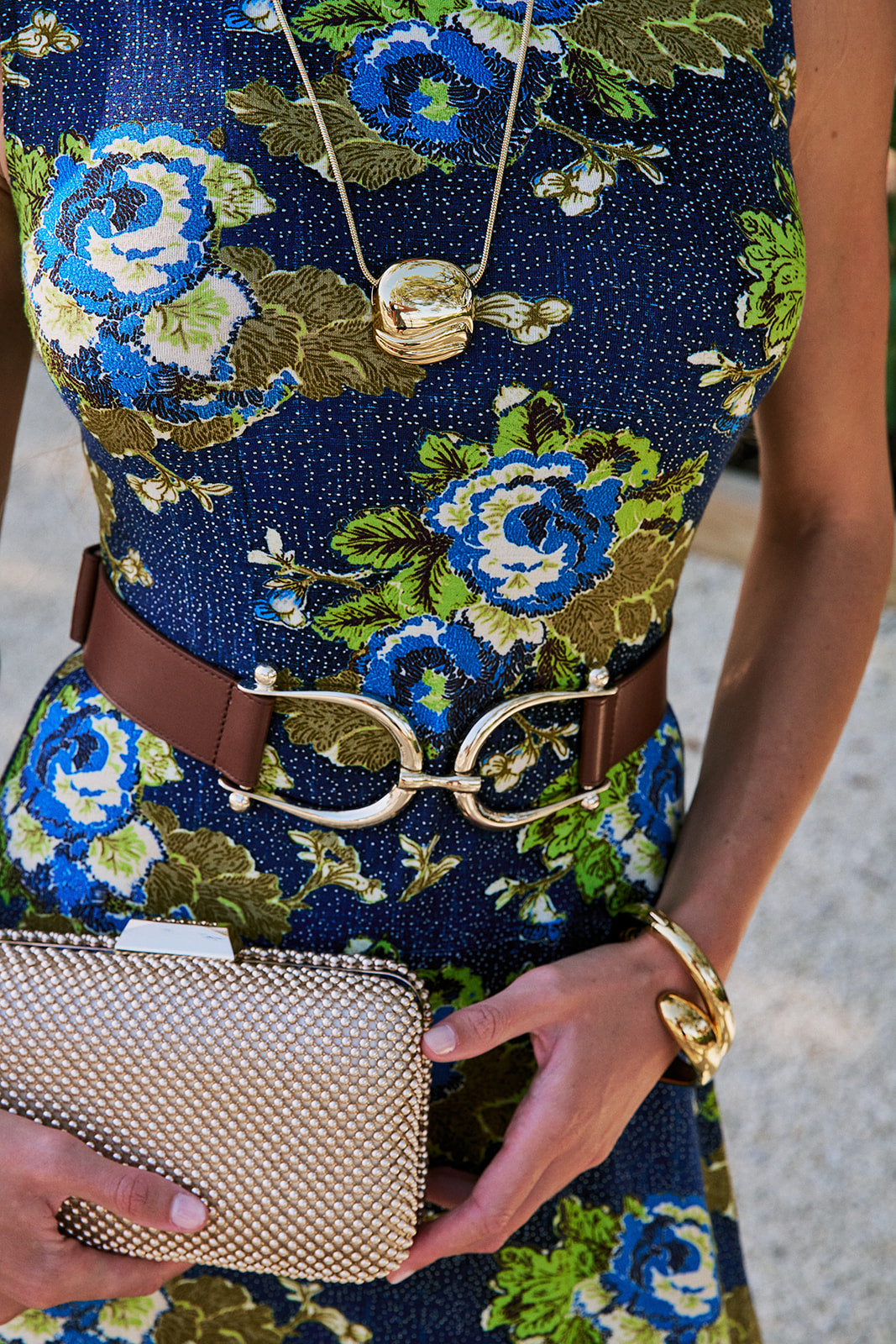 A close-up of a person wearing a navy blue floral dress with blue, green, and white flowers, Grant Belt, holding a gold studded clutch, and wearing gold jewelry.