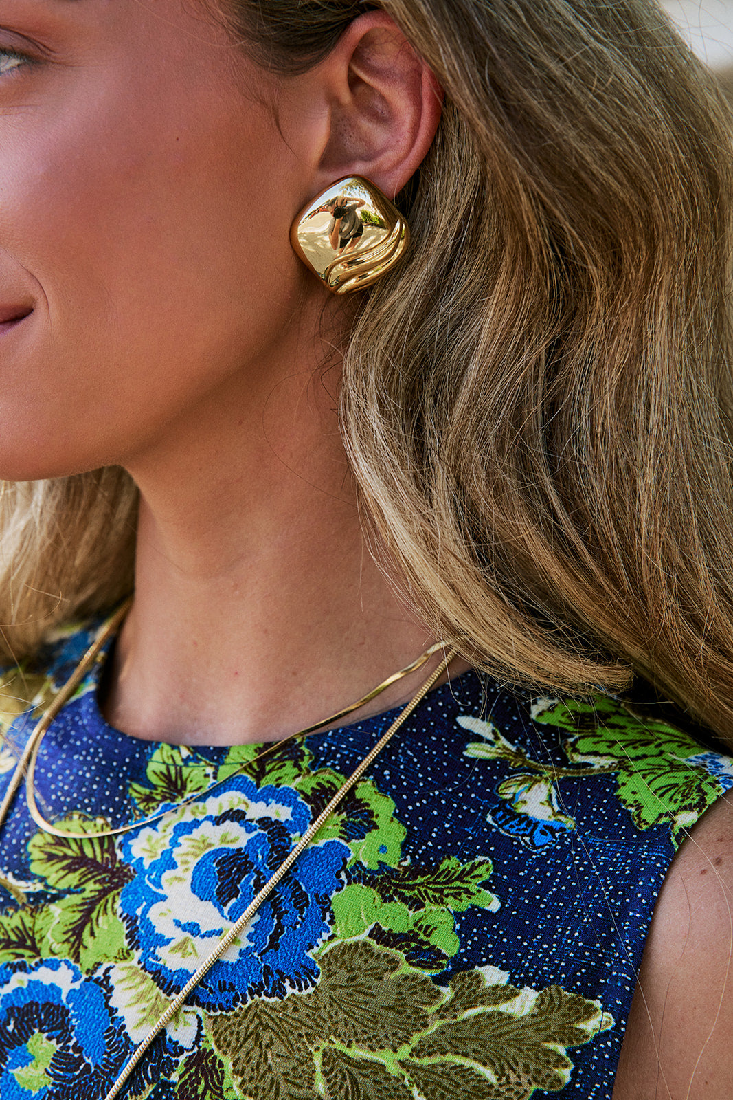 A close-up lifestyle shot of a woman wearing a chunky, organic-shaped gold stud earring, gold necklaces, and a blue floral dress.