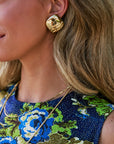 A close-up lifestyle shot of a woman wearing a chunky, organic-shaped gold stud earring, gold necklaces, and a blue floral dress.