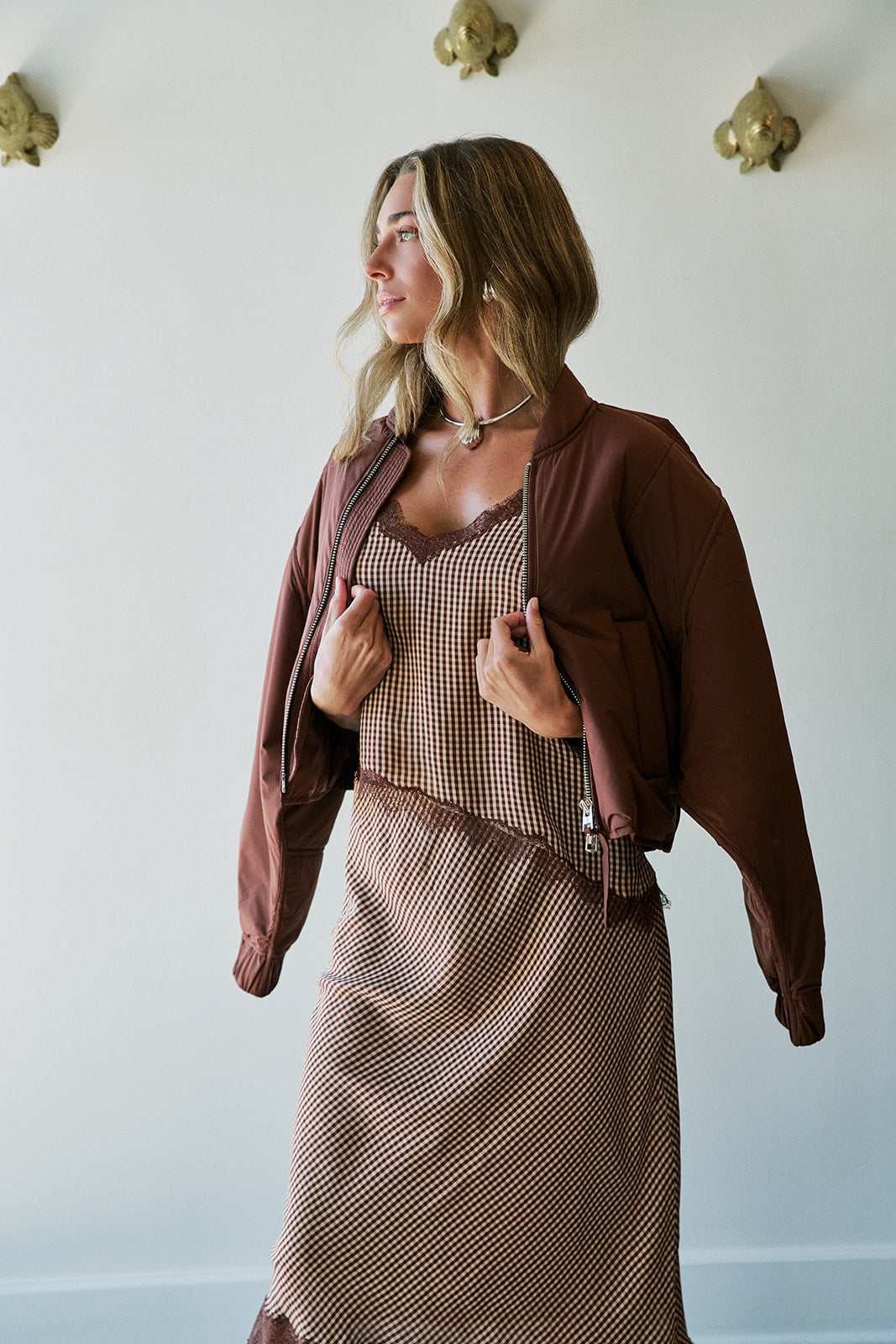 A woman wearing a brown patterned midi set with a matching brown open-front jacket, standing against a white wall with decorative gold wall hooks.