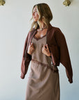 A woman wearing a brown patterned midi set with a matching brown open-front jacket, standing against a white wall with decorative gold wall hooks.