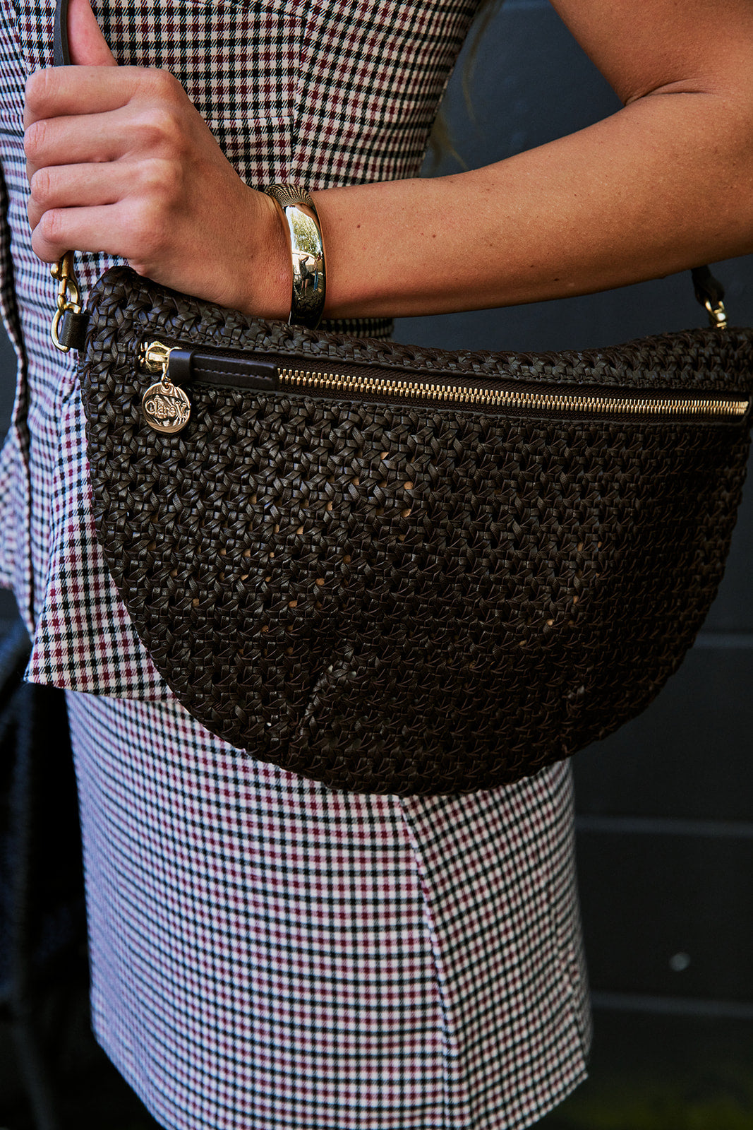 A close-up of a person wearing a dark brown woven faux-leather half-moon shoulder bag with gold-tone hardware, paired with a red, white, and black gingham set.