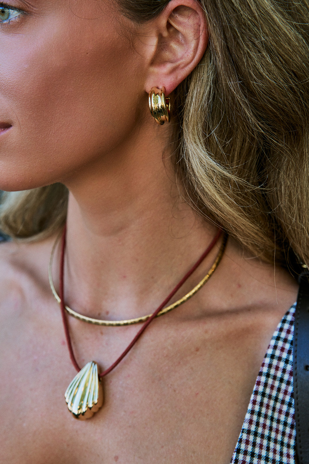 Close-up of a woman wearing layered necklaces and a gold hoop earring. The necklaces include a gold chain and a red cord necklace with a gold seashell pendant.