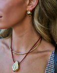 Close-up of a woman wearing layered necklaces and a gold hoop earring. The necklaces include a gold chain and a red cord necklace with a gold seashell pendant.