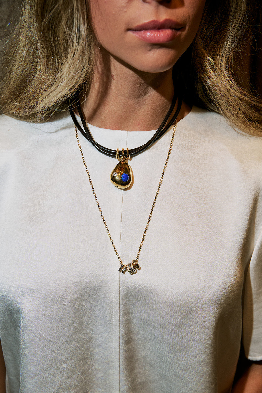 A close-up of a woman wearing layered necklaces, including a black cord choker with a gold teardrop pendant containing blue and white stones, and a delicate gold chain necklace with small gold charms.
