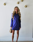 A woman stands indoors wearing a fitted royal blue mini dress with long sleeves and a high neckline, paired with beige strappy heels and a small beige handbag. The background is a white wall with decorative gold wall pieces and light wood flooring.