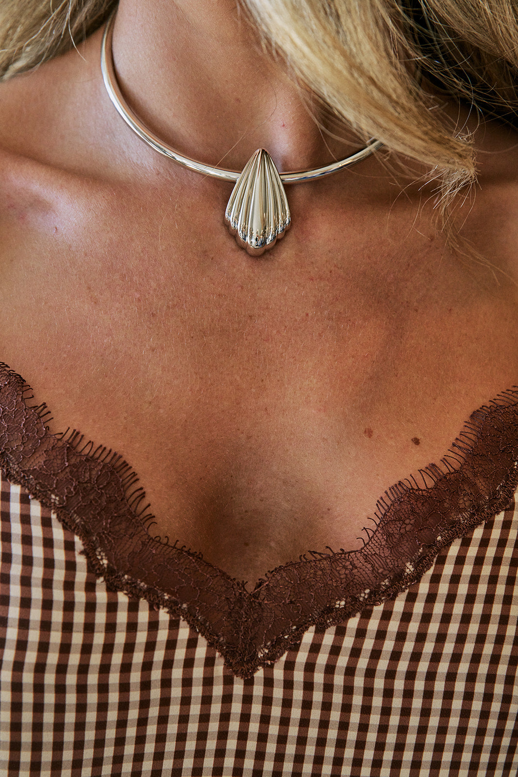 A close-up of a woman's neck and chest wearing a silver choker necklace with a ribbed teardrop pendant, paired with a brown and beige gingham top with lace trim.