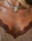 A close-up of a woman's neck and chest wearing a silver choker necklace with a ribbed teardrop pendant, paired with a brown and beige gingham top with lace trim.