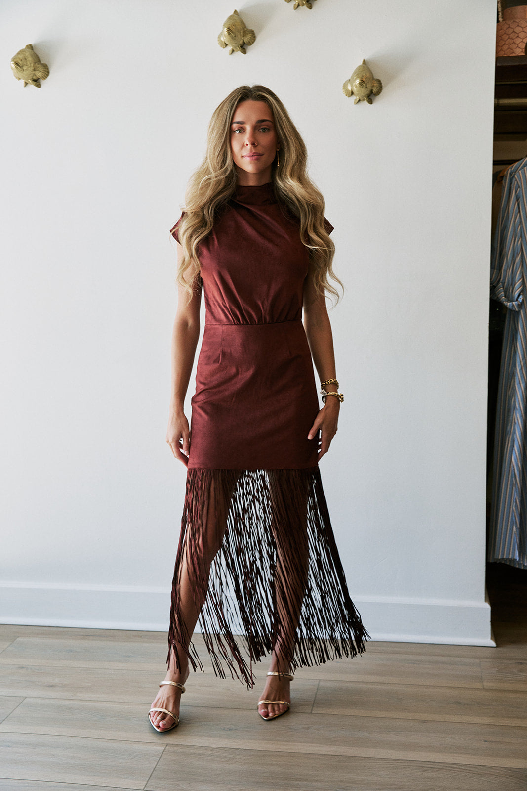 A woman models a sleeveless high-neck dress with a solid rust-brown bodice and a long, flowing black fringe skirt, standing in a room with light wood floors and a white wall.