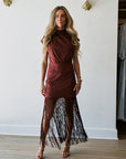 A woman models a sleeveless high-neck dress with a solid rust-brown bodice and a long, flowing black fringe skirt, standing in a room with light wood floors and a white wall.