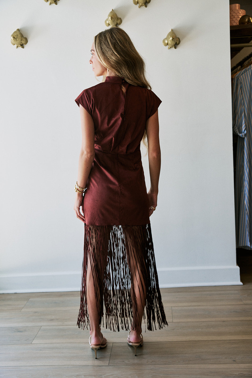 A woman is shown from behind wearing a short-sleeved burgundy dress with a long fringe skirt, standing in a room with a white wall and wood floor.