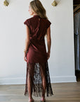A woman is shown from behind wearing a short-sleeved burgundy dress with a long fringe skirt, standing in a room with a white wall and wood floor.