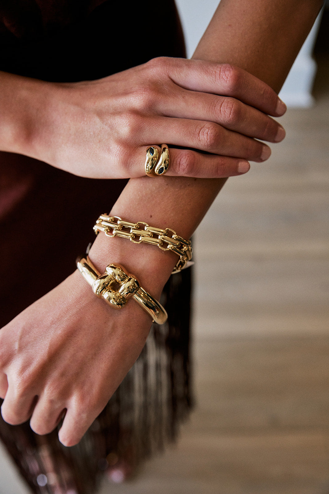 A close-up of a person's hands and wrists wearing multiple gold-toned jewelry pieces, including a snake-shaped ring and chain-link bracelets, against a softly blurred indoor background.
