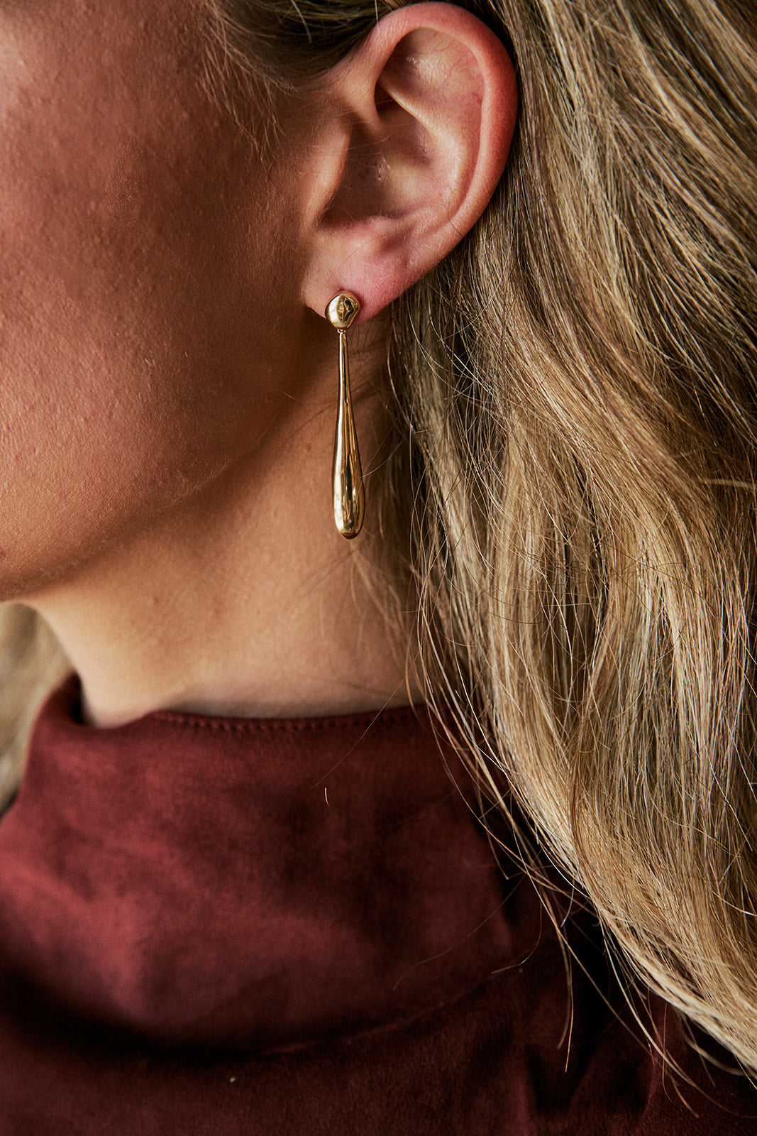 A close-up side profile of a woman wearing a delicate gold teardrop dangle earring