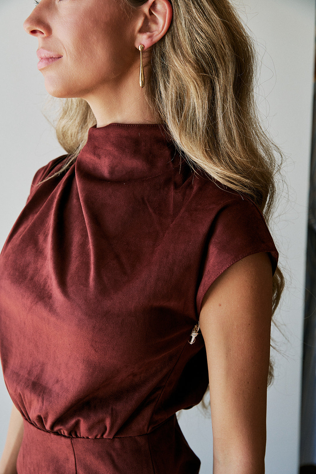 A close-up side profile of a woman wearing a high-neck, short-sleeved burgundy dress with a gathered waist, paired with a gold dangle earring.