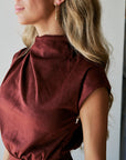 A close-up side profile of a woman wearing a high-neck, short-sleeved burgundy dress with a gathered waist, paired with a gold dangle earring.