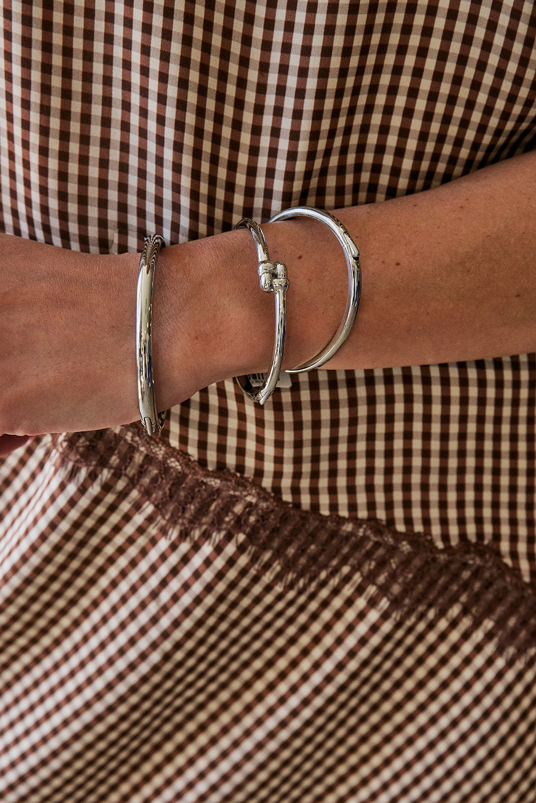 A close-up of a person's wrist wearing three silver-toned bangles, including a textured flat band, a small bead-joined loop, and a larger open-ended bangle, set against a brown and beige gingham patterned garment.