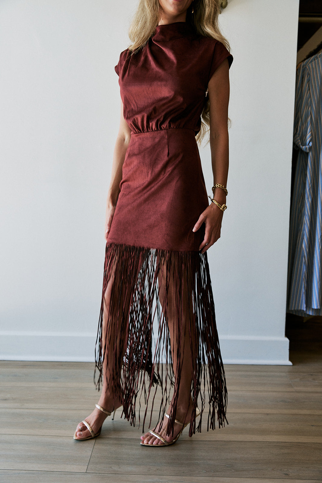 A woman wearing a sleeveless rust-brown mini dress with a gathered waist and a long fringe hem, paired with clear high-heeled sandals, standing in a room with light wood floors and a white wall. Front view