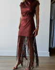 A woman wearing a sleeveless rust-brown mini dress with a gathered waist and a long fringe hem, paired with clear high-heeled sandals, standing in a room with light wood floors and a white wall. Front view