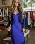 A woman stands inside a clothing boutique wearing a long royal blue satin slip dress with a matching cropped jacket and Luna Nera Pendant Necklace. She is holding a small gold-colored handbag, with racks of clothing and a window visible in the background.