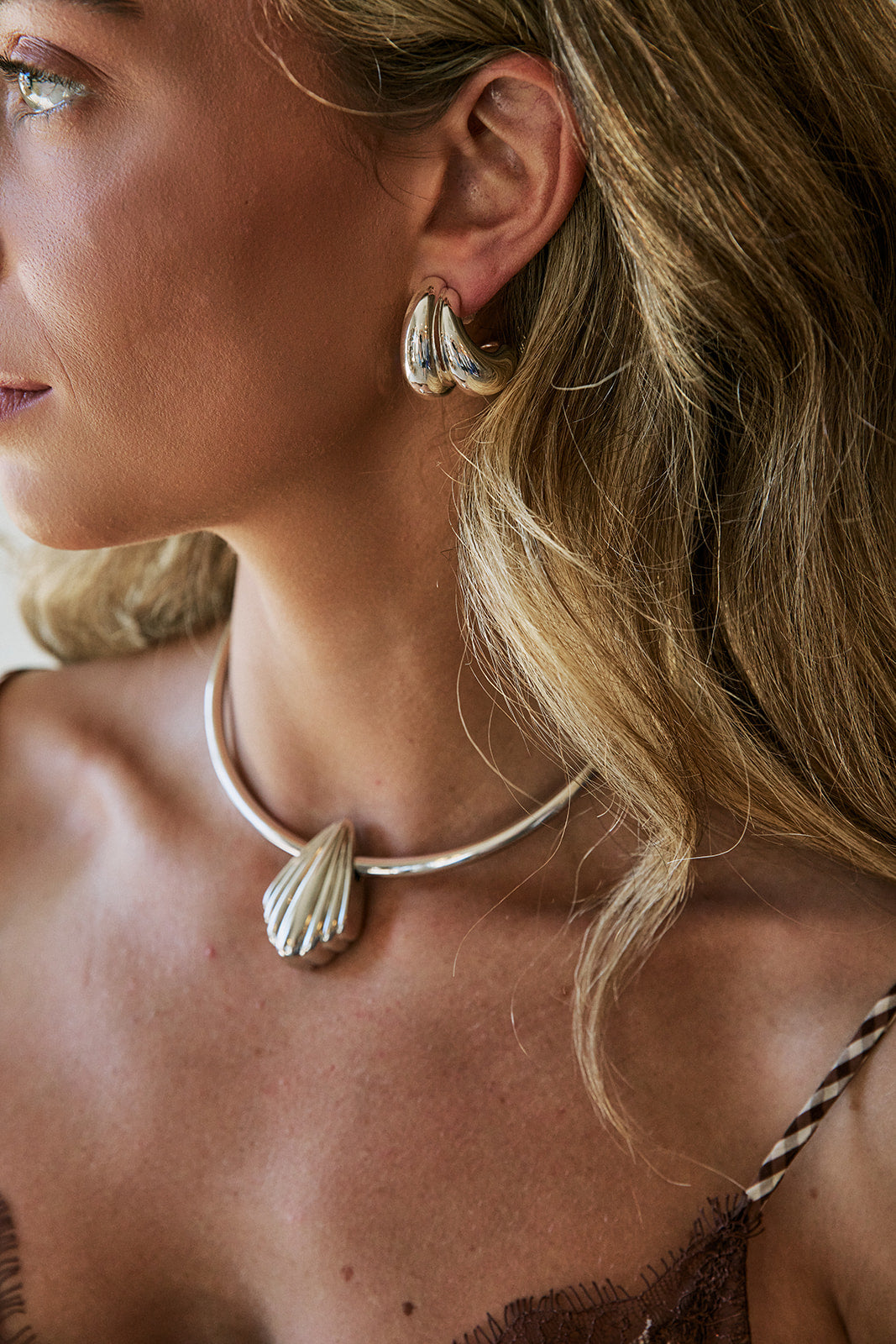 A close-up lifestyle shot of a woman wearing a silver shell-shaped choker necklace and matching silver huggie hoop earrings.