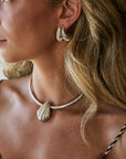 A close-up lifestyle shot of a woman wearing a silver shell-shaped choker necklace and matching silver huggie hoop earrings.
